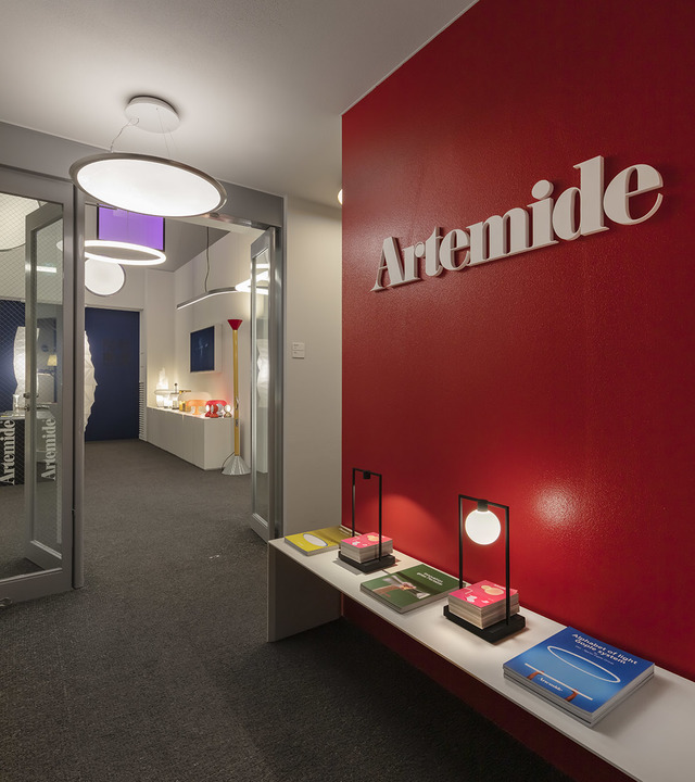 Artemide showroom entrance with red branded wall, table lamps with globe shades on shelf, and circular pendant lamps in corridor