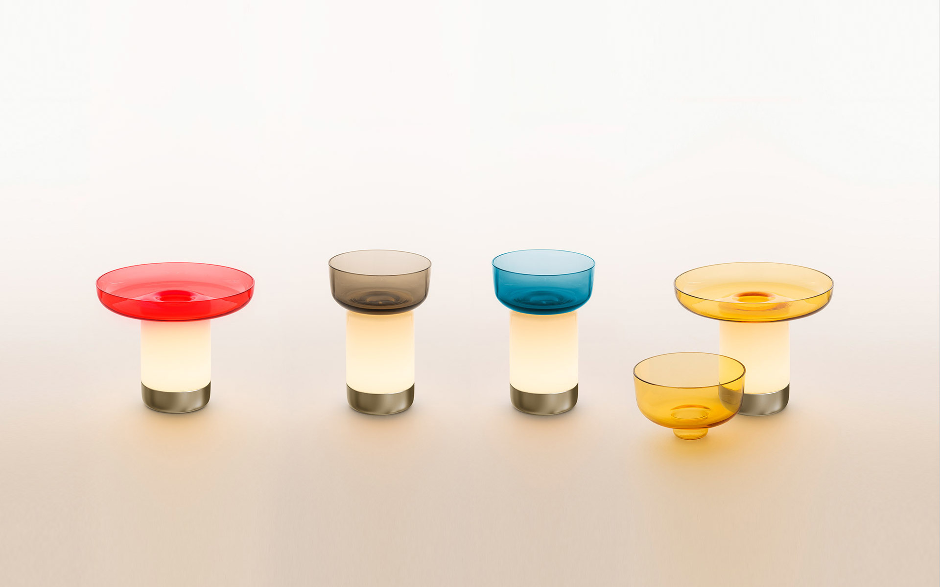 Four Artemide portable lamps with glowing white bases, metal feet, and colored glass bowls in red, smoke, teal, and amber