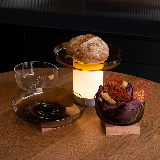 Artemide Unplugged portable lamp with warm white glow on a wooden table, surrounded by glass bowls with bread and snacks