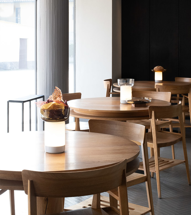 Restaurant dining area with round wooden tables and chairs, featuring Artemide portable table lamps with white cylindrical shades providing warm ambient light.