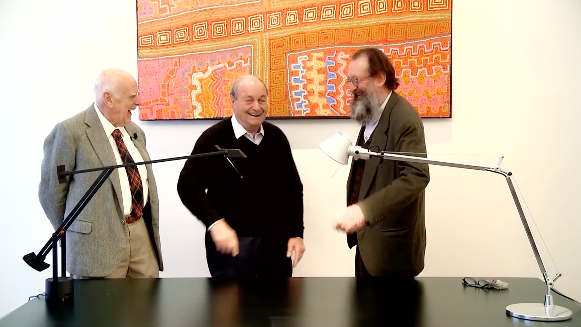 Three men laughing together in a bright room, flanked by two Artemide Tolomeo desk lamps on a dark table, colorful artwork behind.