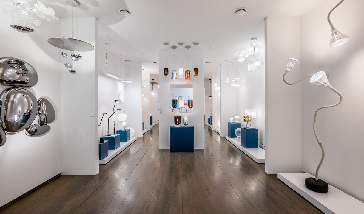 Artemide showroom with white walls, dark wood floor, displaying pendant, table, and floor lamps on blue pedestals