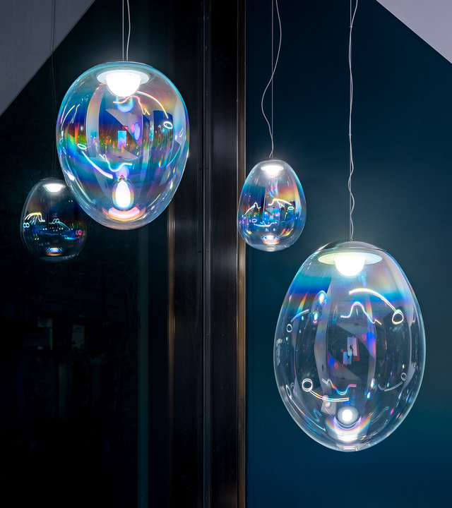 Four iridescent glass pendant lamps in varying oval sizes, reflecting rainbow hues against a dark teal background.