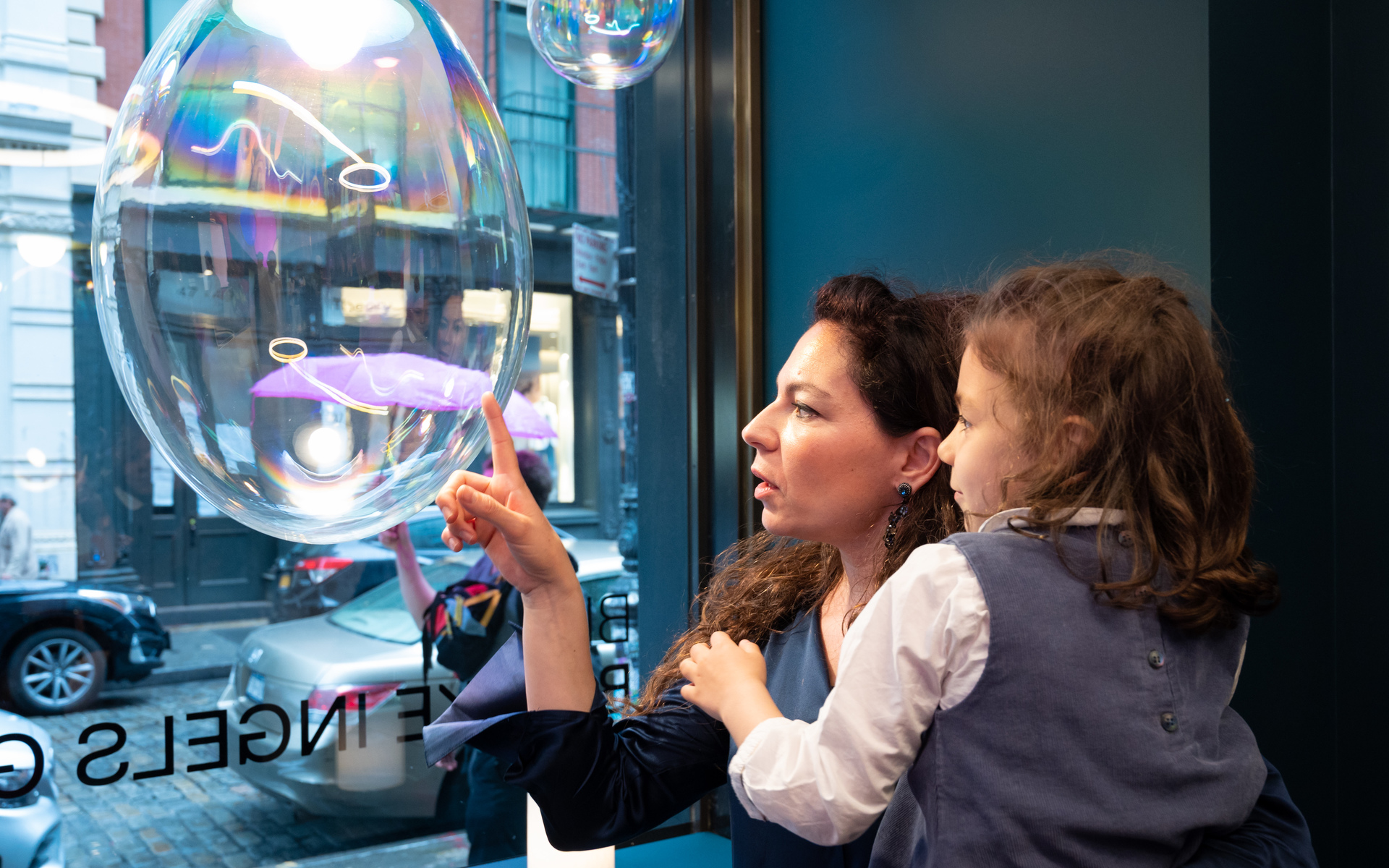 Woman holding a child, both fascinated by a large soap bubble near a shop window with city street in background