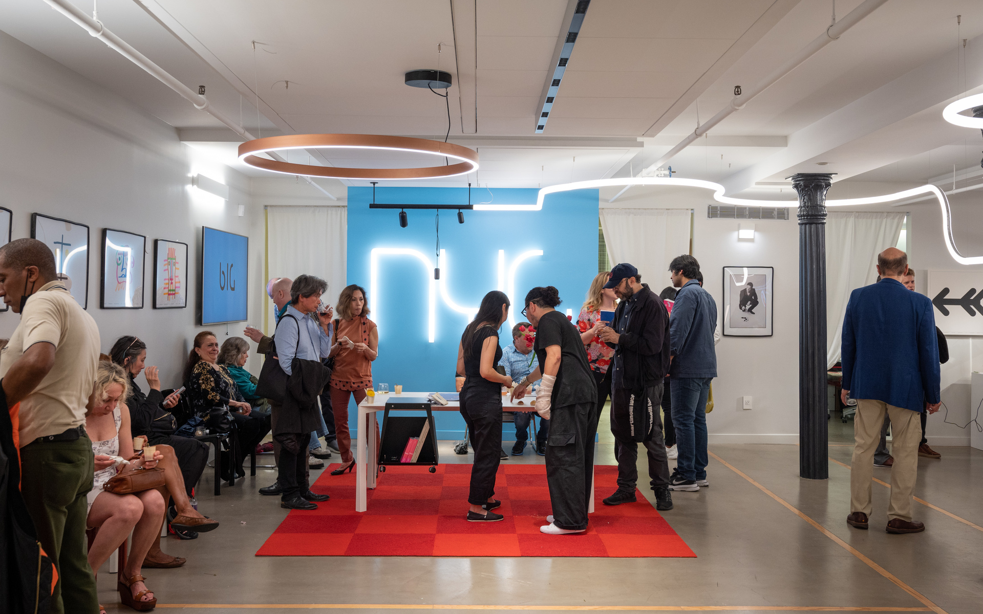 Gallery event with Artemide circular pendant and neon wall art; guests mingle around a table on a red carpet under warm lighting.