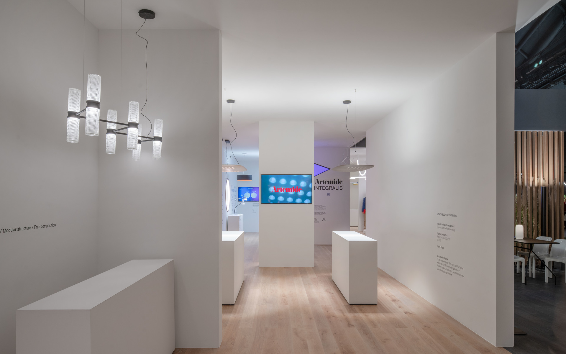 Artemide exhibition stand with a chandelier featuring cylindrical glass diffusers on a horizontal bar, white display plinths, and pendant lights.