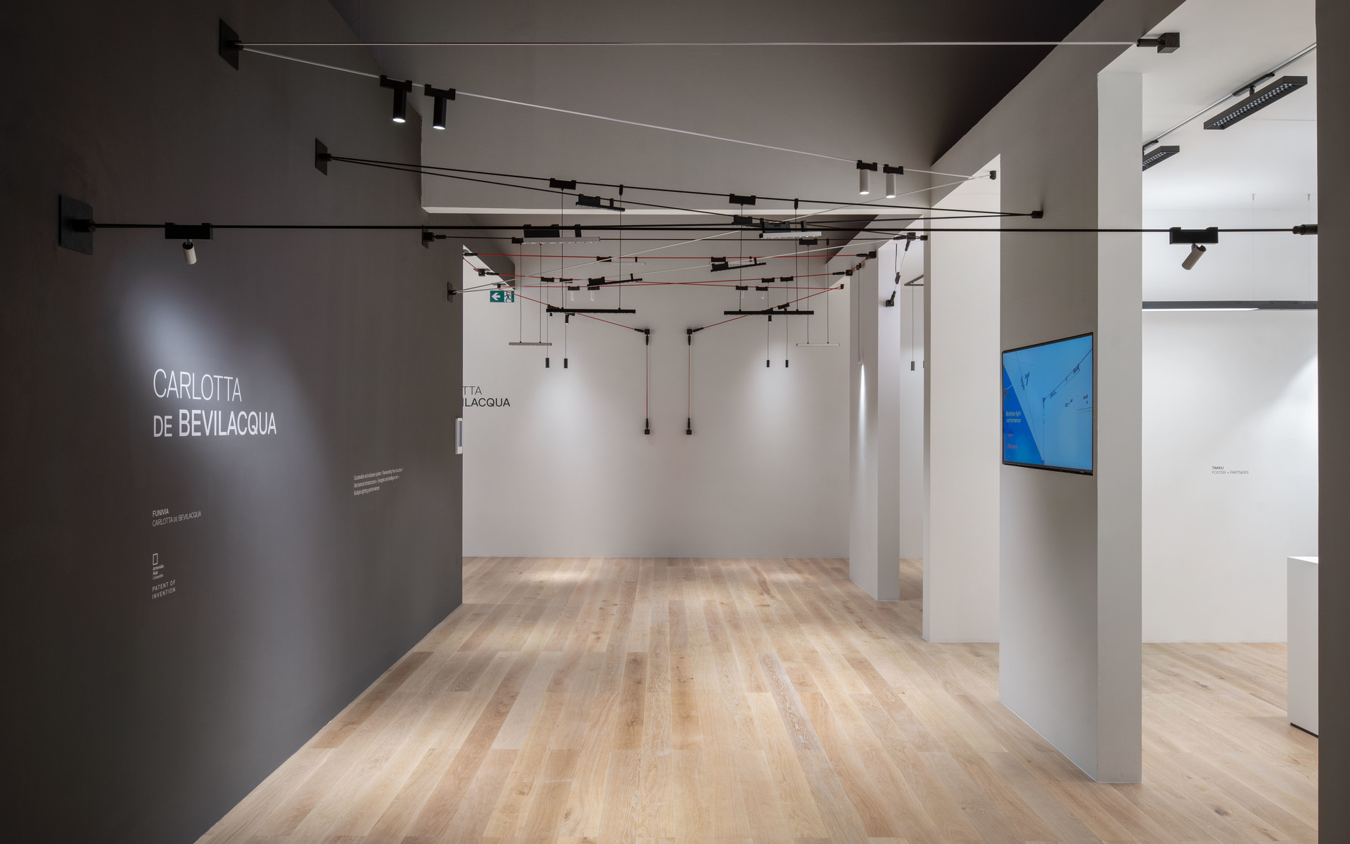 Gallery space with black track lighting system on ceiling, illuminating white walls and light wood floor. Text reads 'Carlotta De Bevilacqua'.