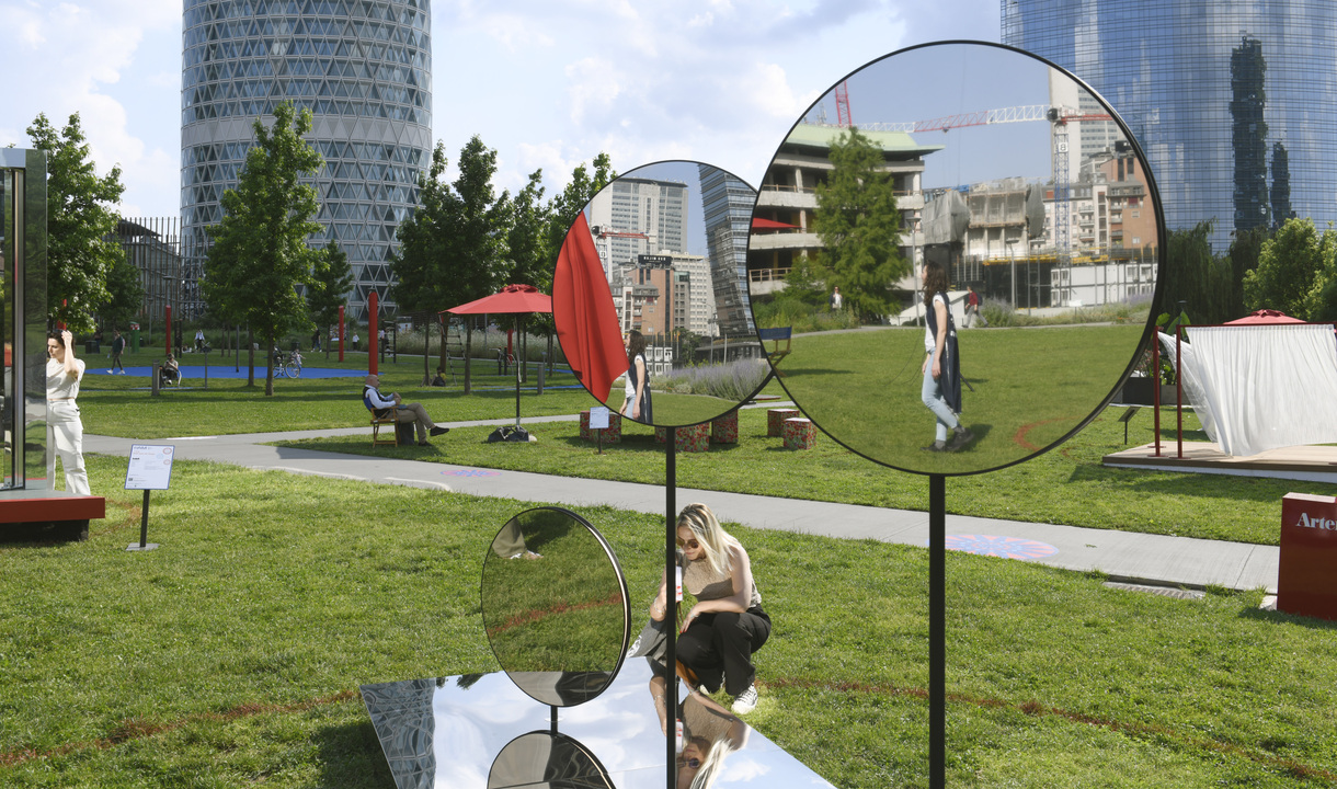 Artemide outdoor installation with circular mirrors on stands in an urban park, reflecting visitors and city skyline with towers.