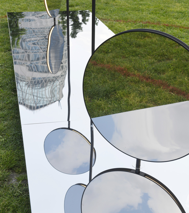 Mirrored circular and rectangular panels on black metal stands outdoors on grass, reflecting sky and surroundings.