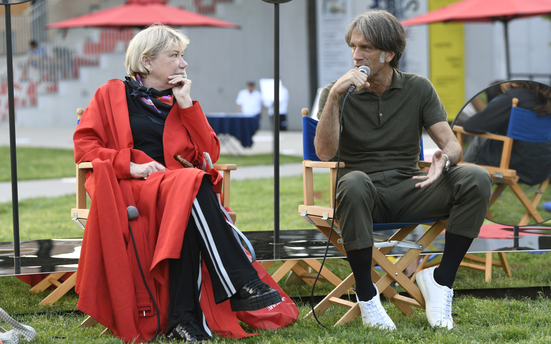 Two people seated in director's chairs outdoors, engaged in a conversation, one holding a microphone, on a grass area.