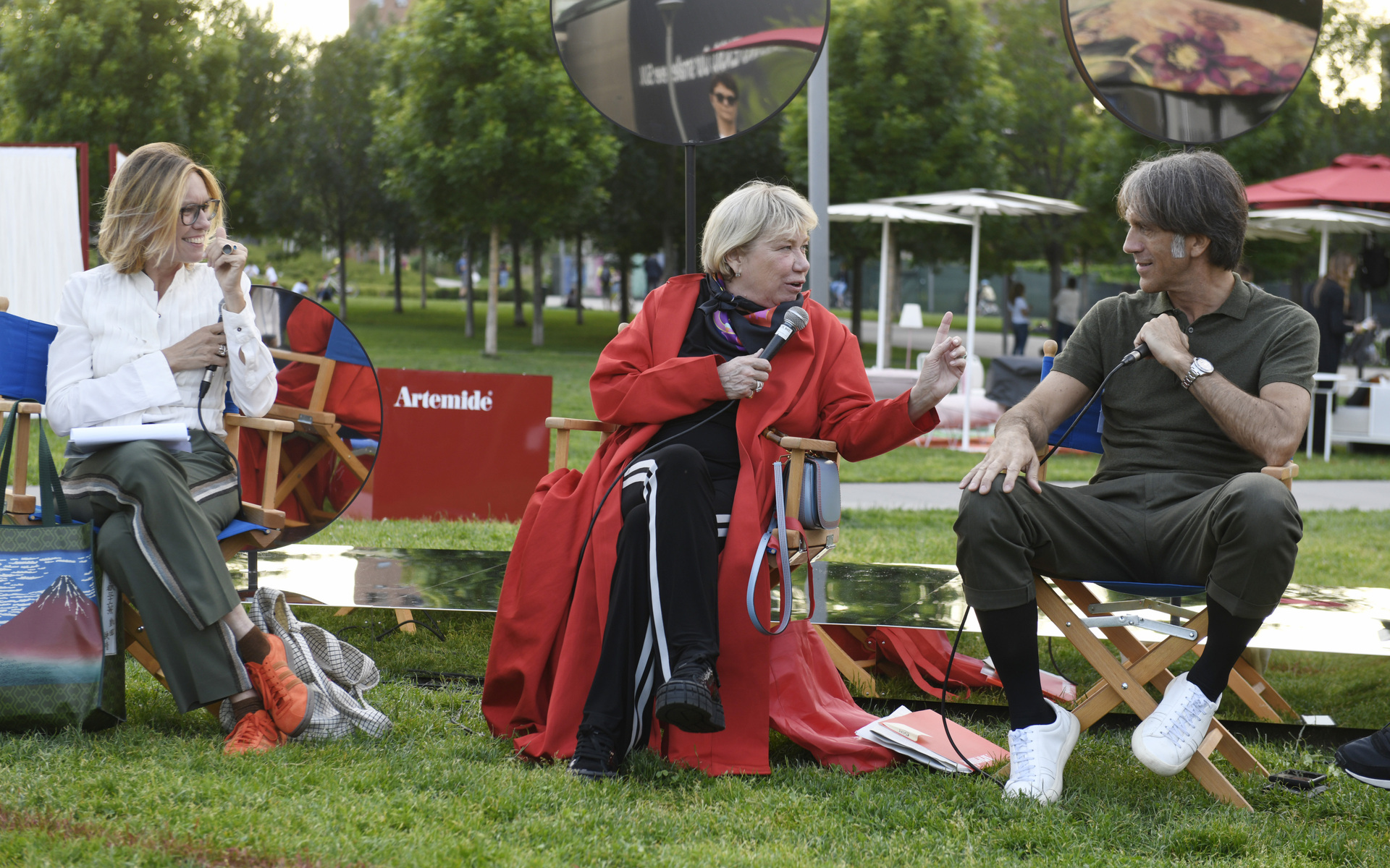 Three people in conversation at an outdoor Artemide event, seated on director's chairs on a lawn, with one speaker holding a microphone.