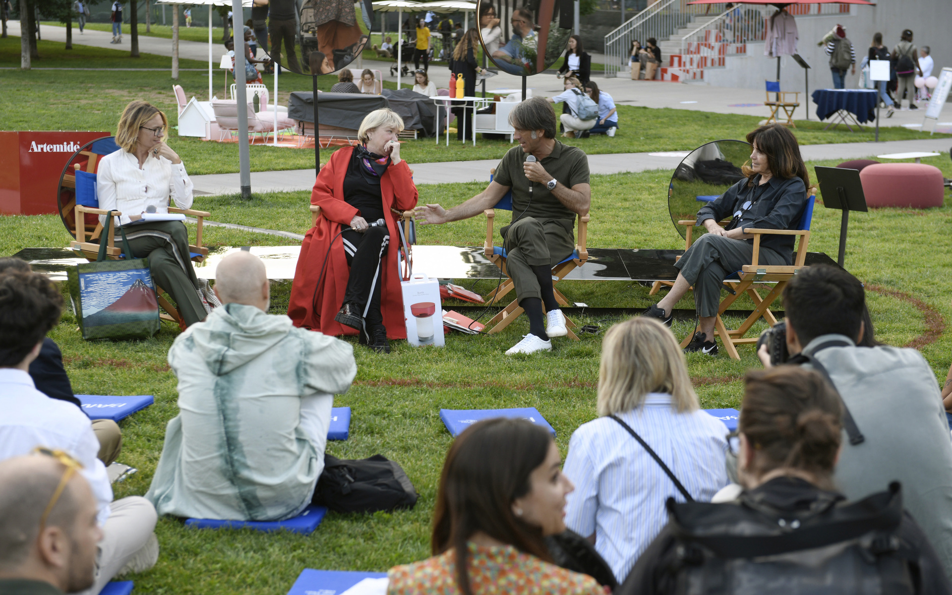 Outdoor panel discussion at an Artemide event, with speakers seated in director chairs on grass, engaging an audience seated on blue mats.