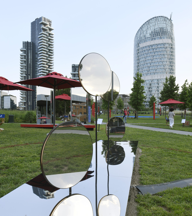 Polished circular mirror sculptures on metal stems in a Milan urban park, with modern skyscrapers and red parasols in background