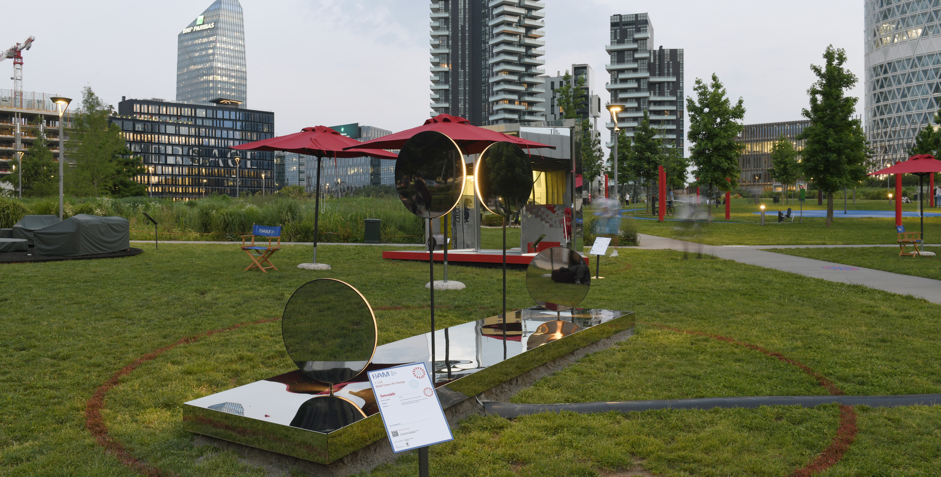 Outdoor art installation with circular mirrors and red accents on grass, set against modern Milan skyscrapers in an urban park.