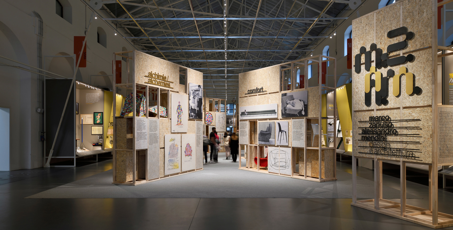 Exhibition hall with plywood display panels showcasing design drawings, photos, and texts, with visitors walking through the corridor.