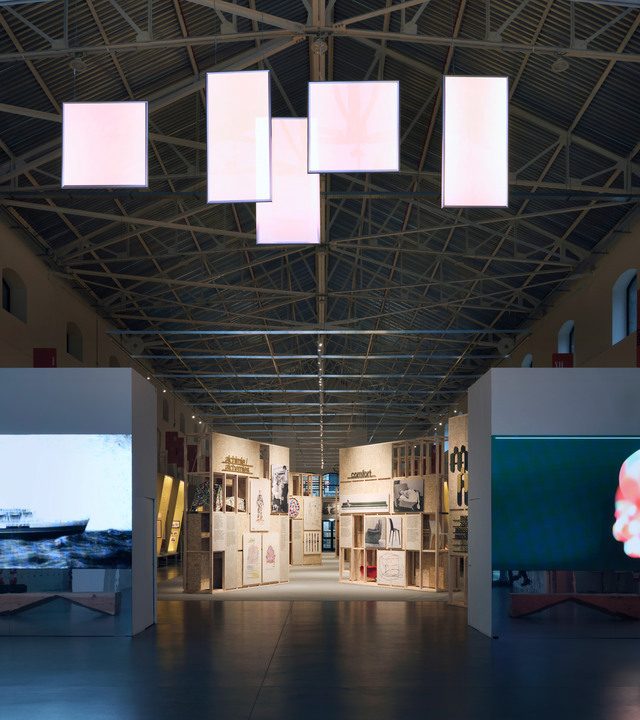 Exhibition hall with large glowing rectangular light panels suspended from industrial truss ceiling, illuminating display corridors below