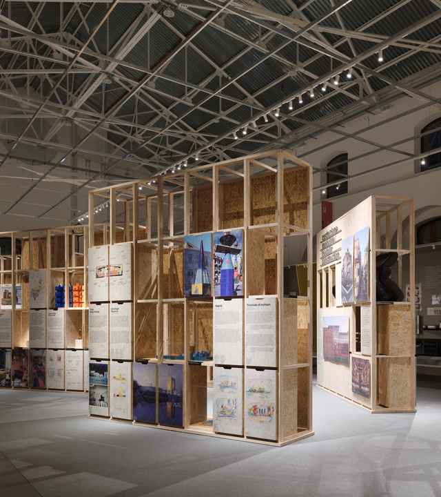 Exhibition space with modular wooden shelving units displaying colorful architectural panels and project boards in an industrial hall.