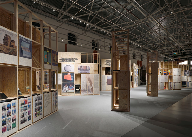 Exhibition hall with wooden modular display structures showing posters and objects, lit by ceiling track spotlights in an industrial space