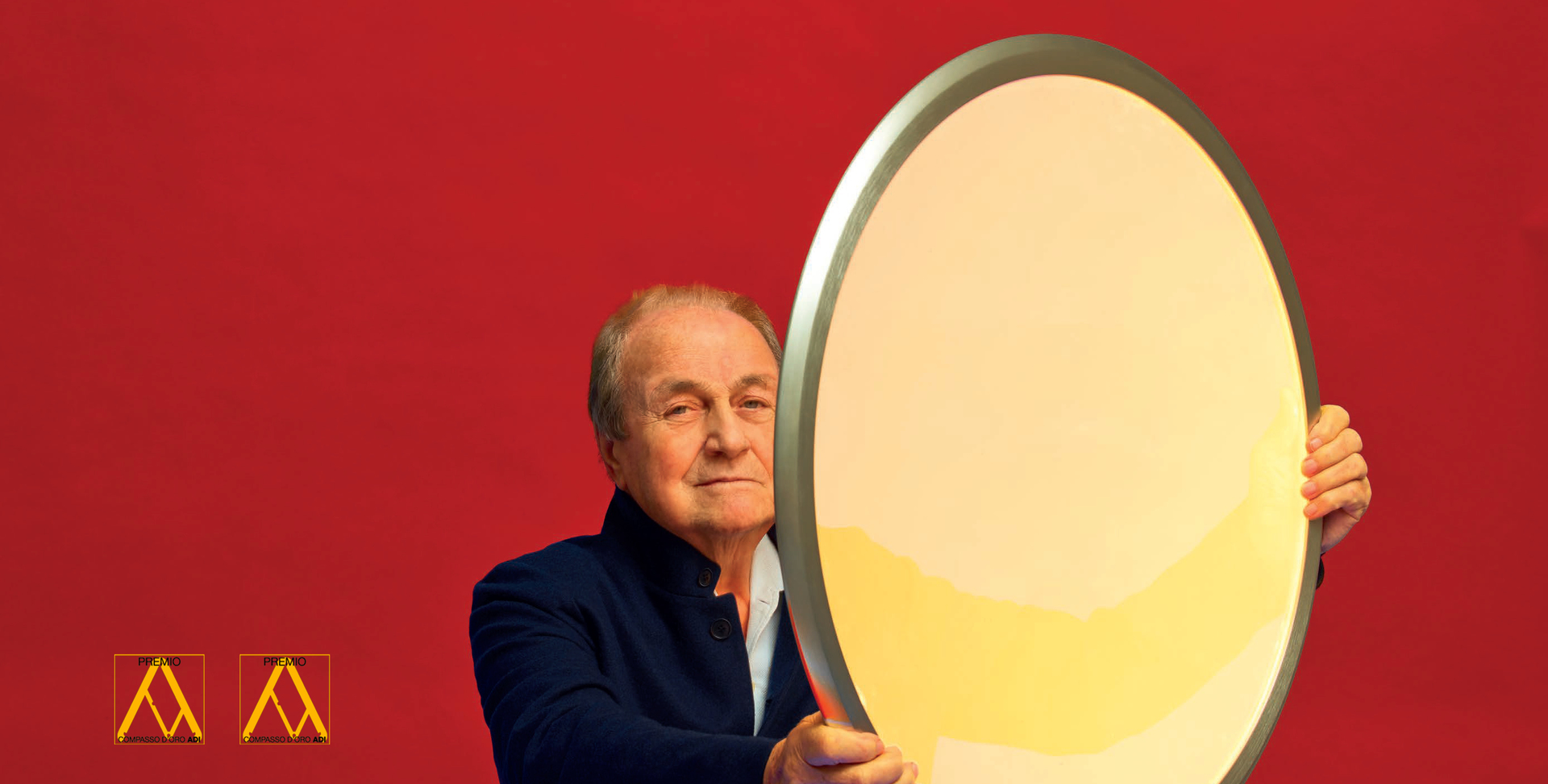 Designer holding a large oval Artemide lamp with white diffuser and silver frame against a red background.
