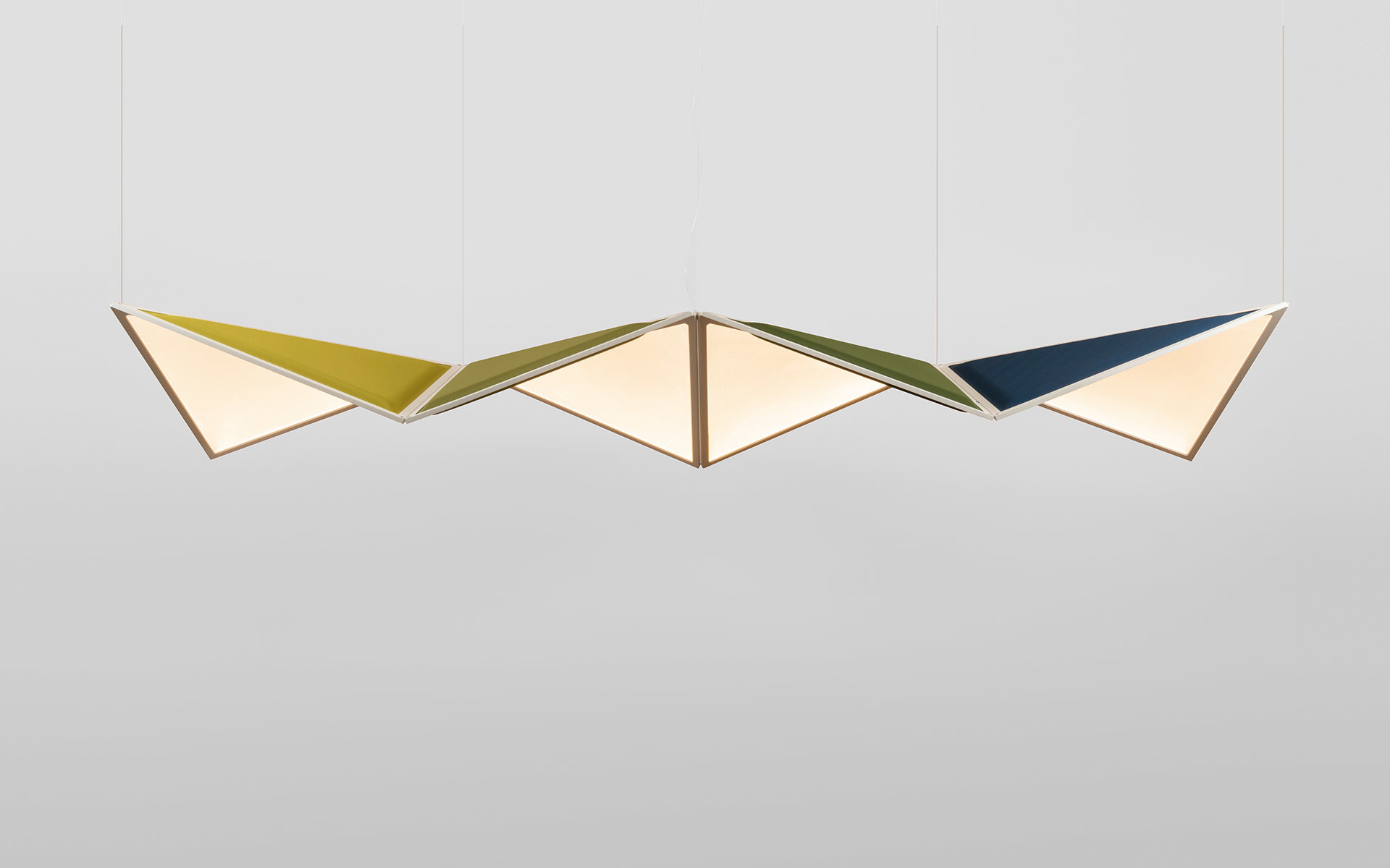 Three geometric diamond-shaped pendant lamps in yellow, olive green, and navy blue, emitting warm light from angular diffusers