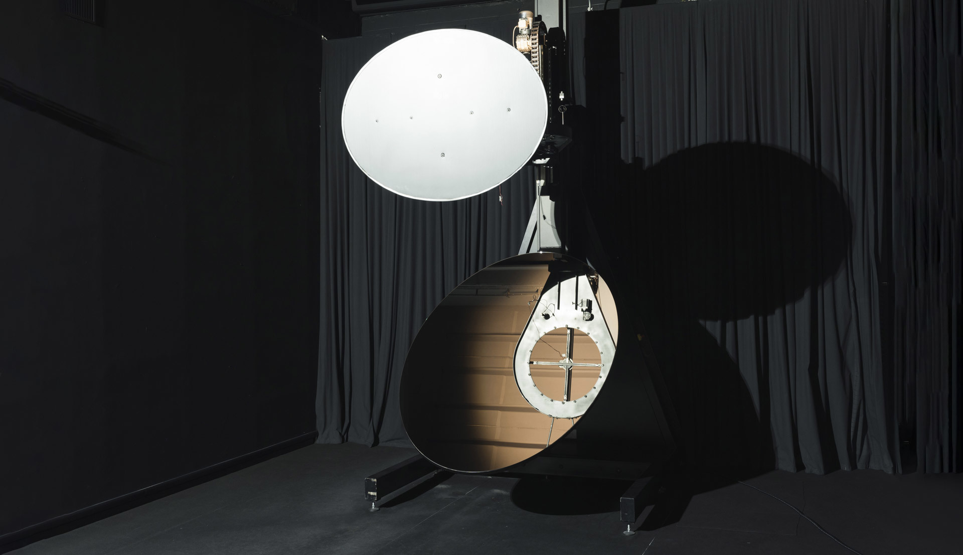 Artemide lamp prototype with large white circular diffuser mounted on a stand next to a wooden egg-shaped structure in a dark studio