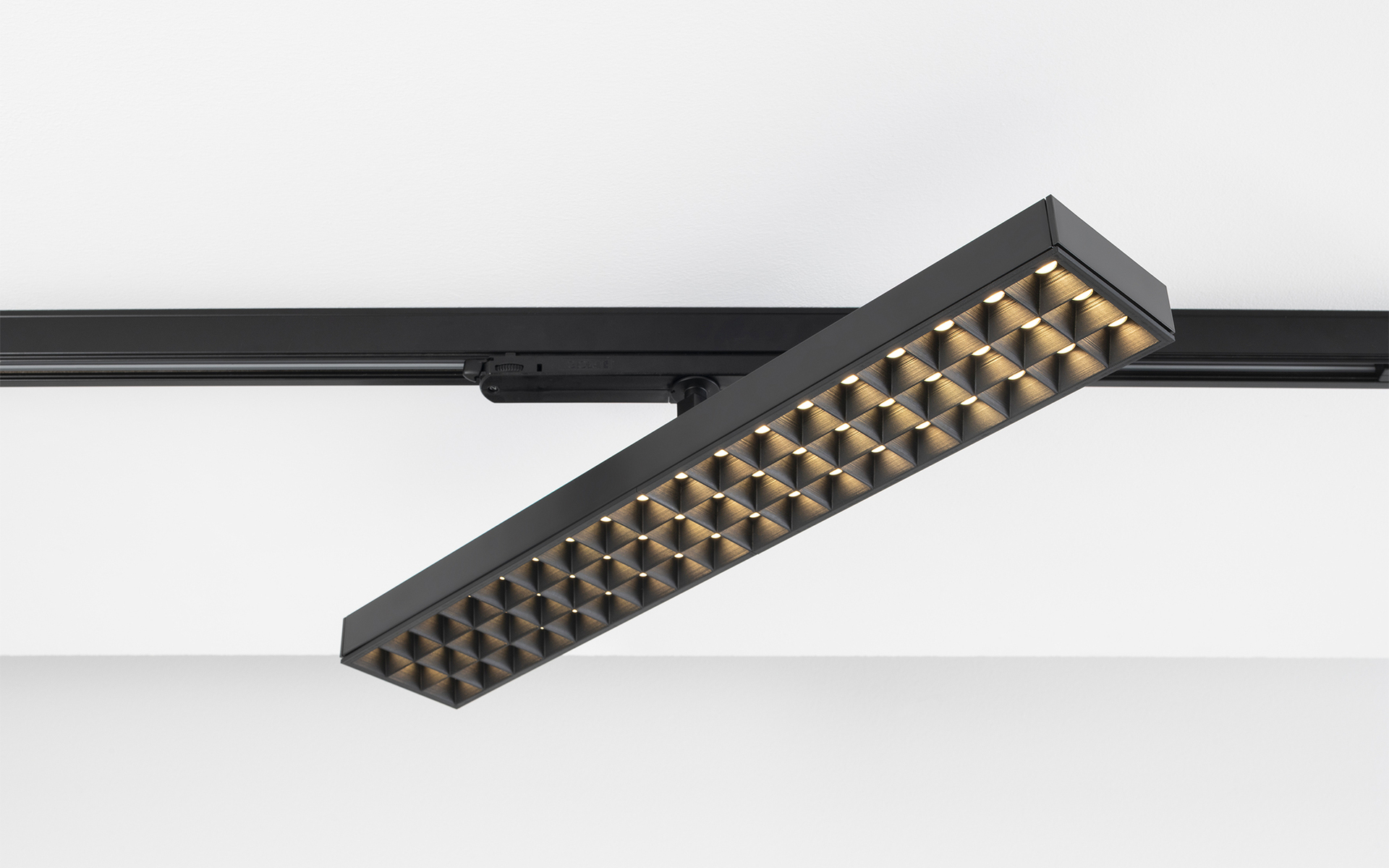 Black rectangular LED track spotlight with honeycomb anti-glare grid, mounted on a black rail system, emitting warm white light