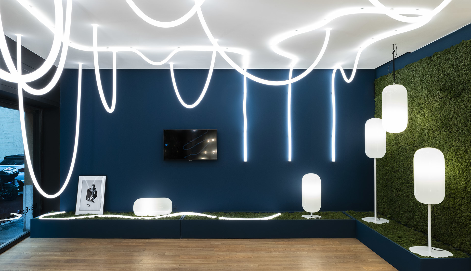 Artemide showroom with curving LED ceiling lights, white oval floor and pendant lamps, against a deep blue wall and green moss panel.