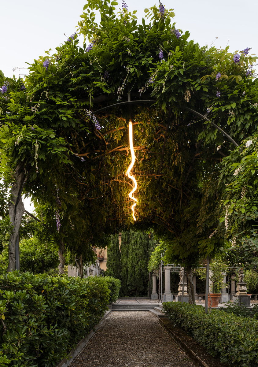 Artemide sinuous glowing light installation hanging from a lush green tree canopy in an elegant garden pathway at dusk