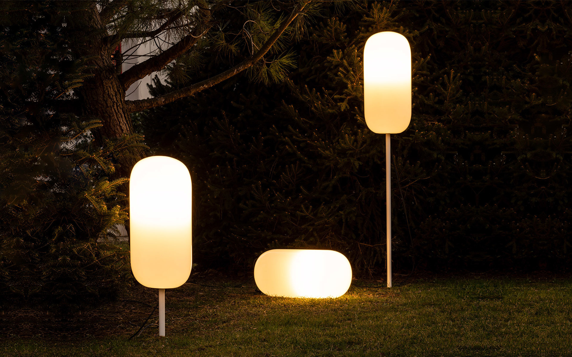 Three Artemide outdoor lamps with glowing white diffusers on a lawn at dusk: two tall pole-mounted and one low ground-level unit.
