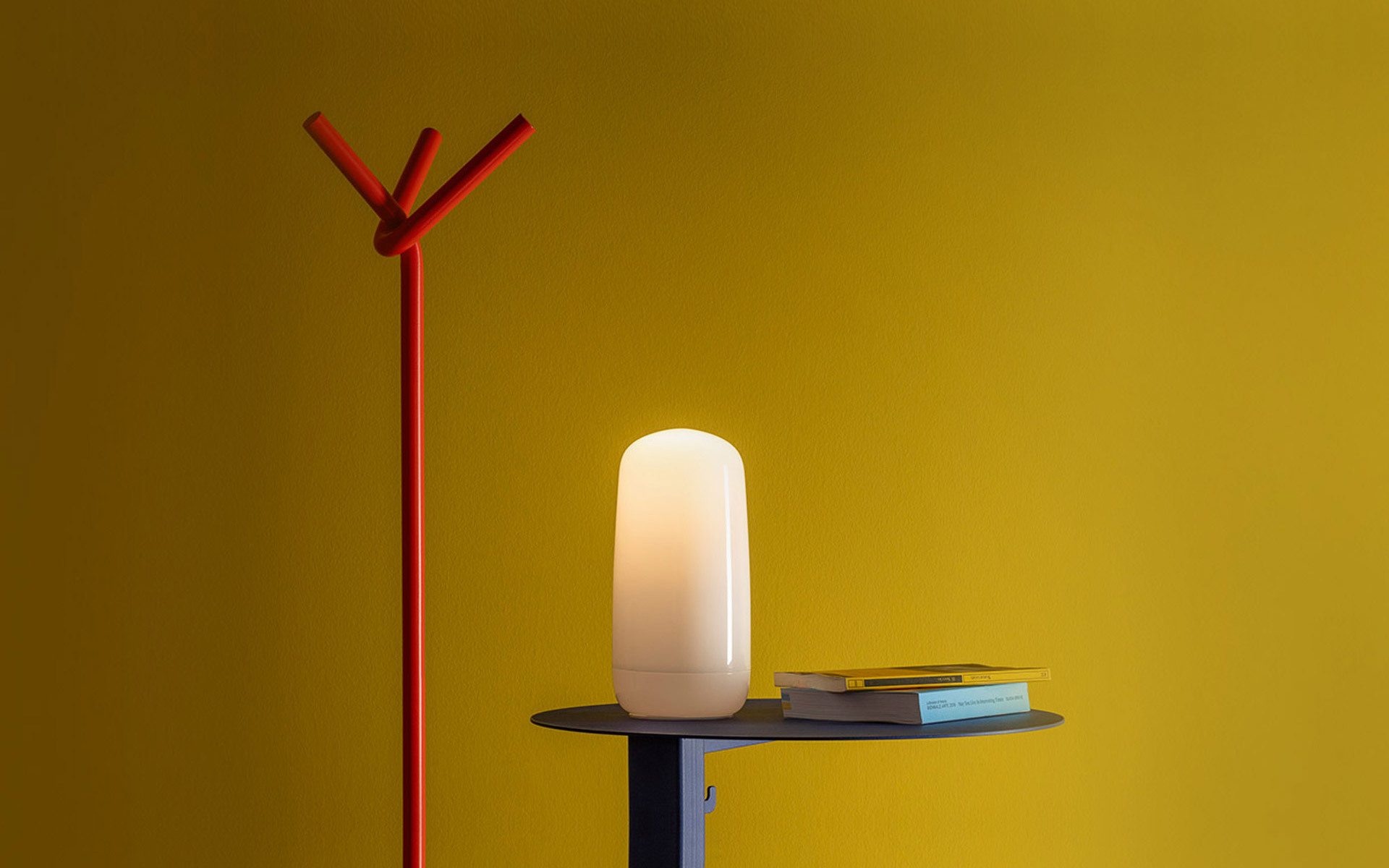 Glowing white oval portable lamp on a dark side table, beside books, against a mustard yellow wall with a red coat stand.