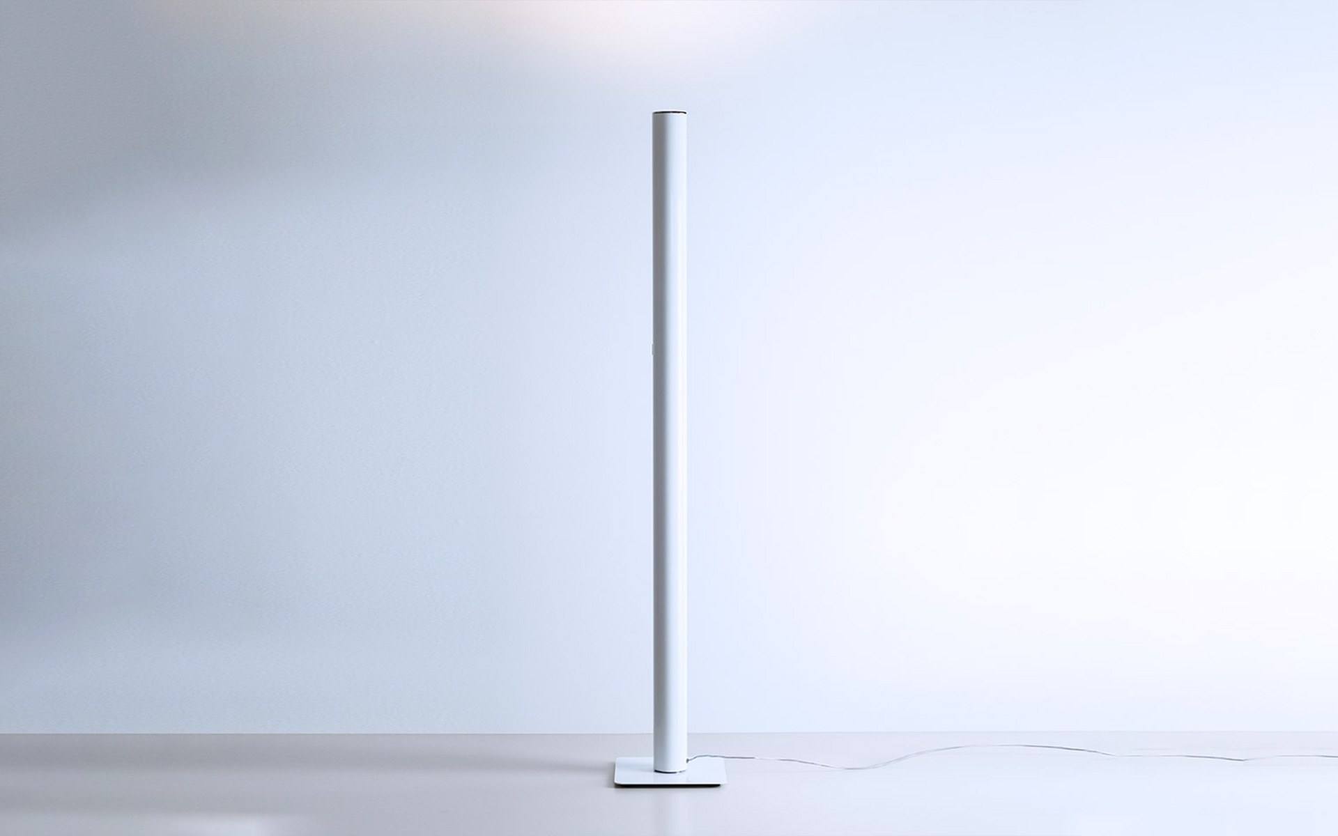 Slim white cylindrical floor lamp on a square base, emitting soft upward light, standing against a light grey wall