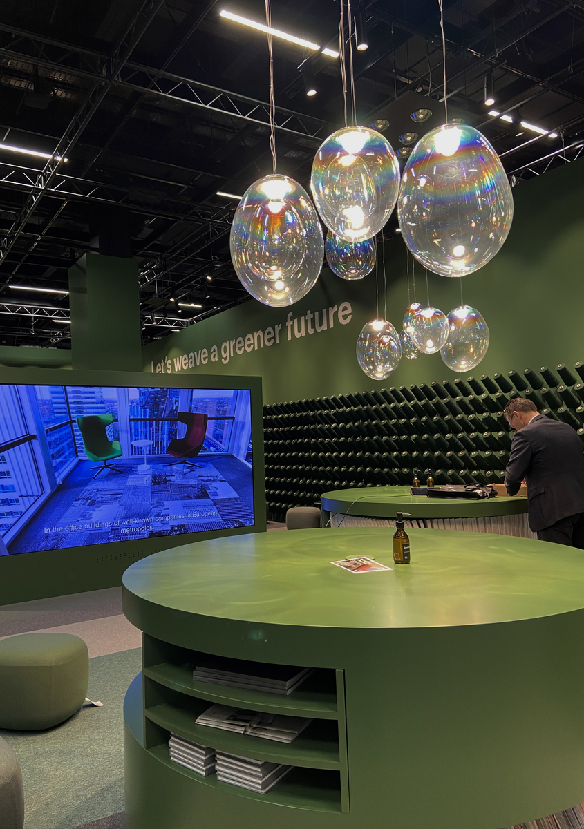 Artemide exhibition stand with iridescent glass bubble pendant lights hanging above a round green table, slogan 'let's weave a greener future'