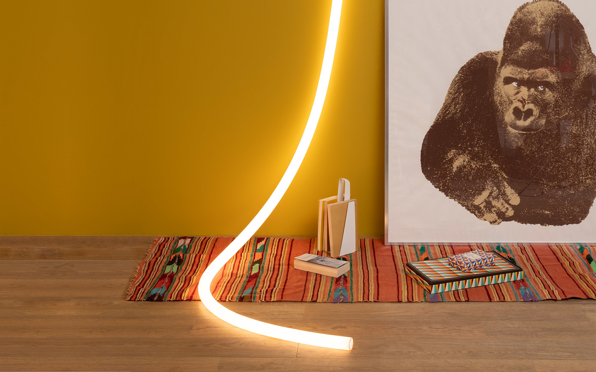 Artemide flexible LED light tube curving from floor to wall, glowing warm white, beside a colorful rug and gorilla artwork