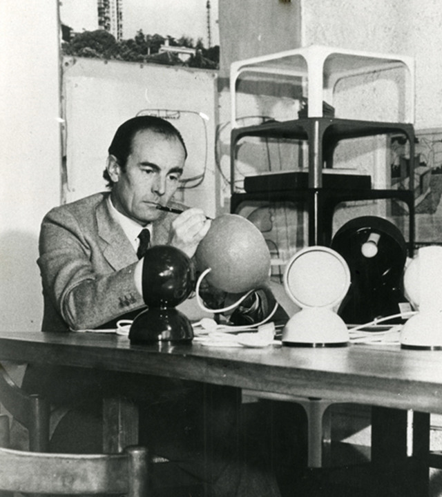 Designer examining spherical lamp prototypes at a workbench, with Artemide Eclisse and other globe-shaped lamps displayed on the table.