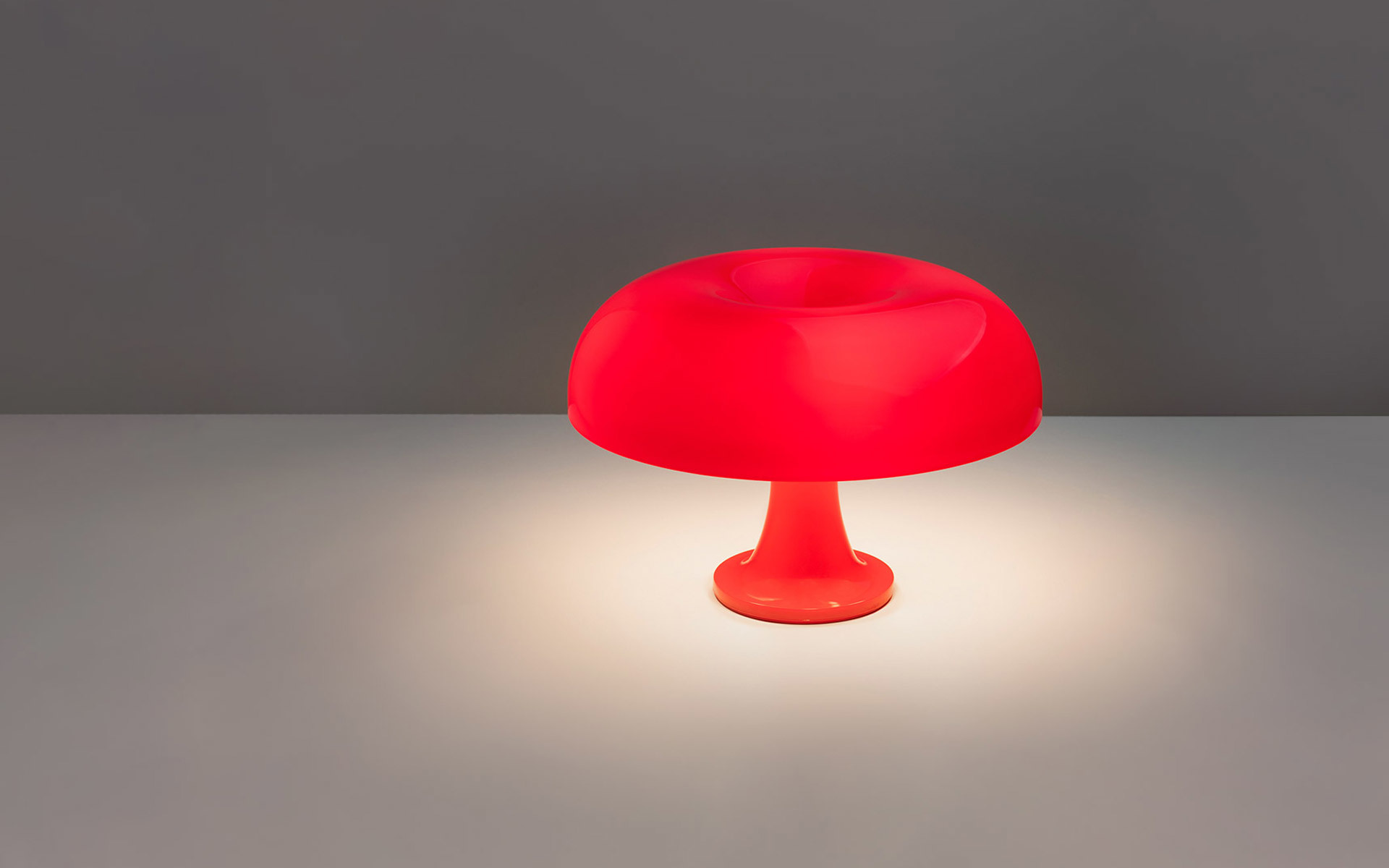 Artemide Nessino table lamp in red, mushroom-shaped diffuser on a slender base, casting warm light on a surface