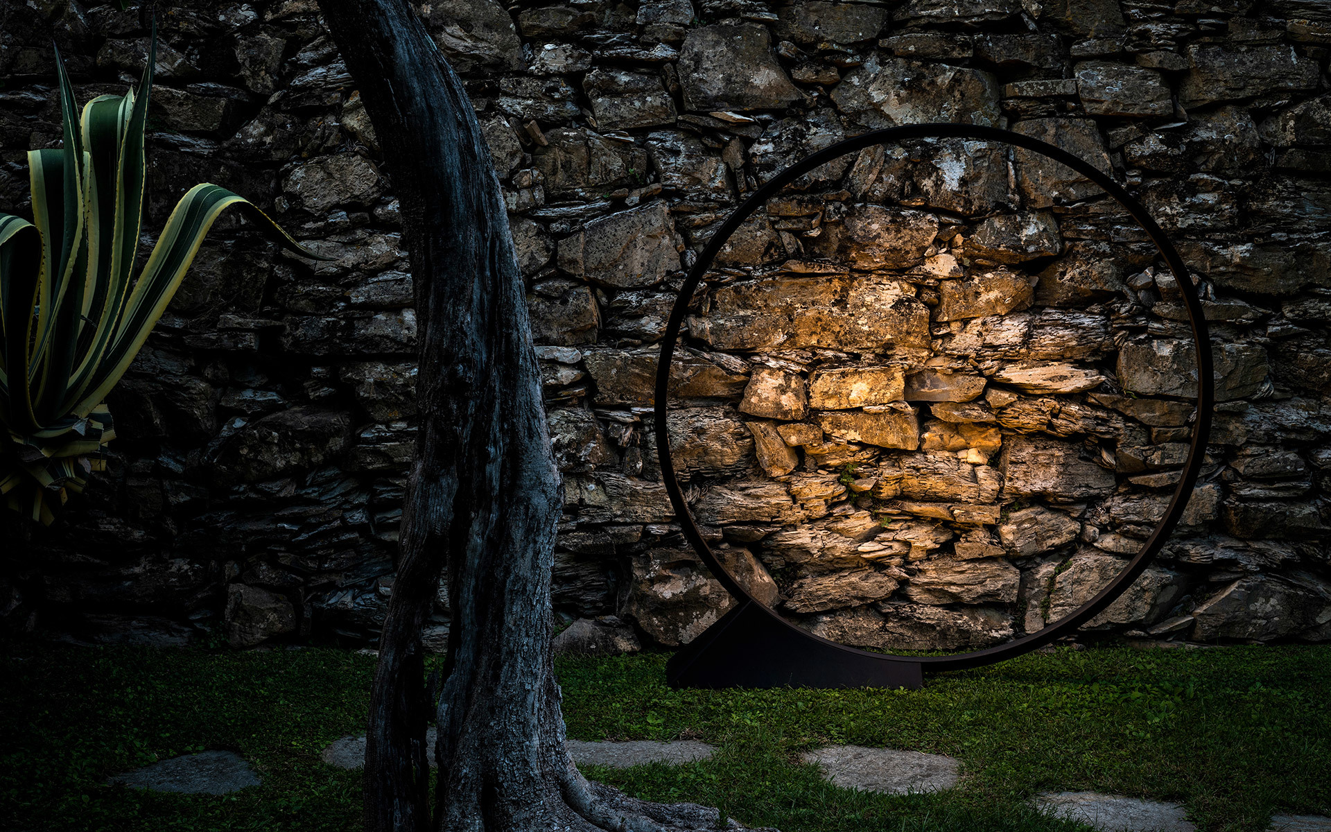 Circular black metal floor lamp illuminating a rustic stone wall in an outdoor garden at night, with agave plant nearby