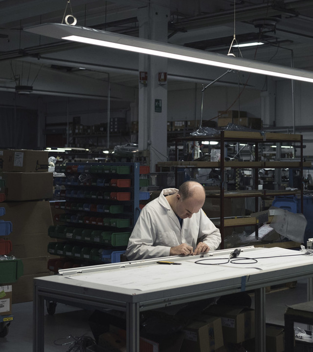 Technician assembling a lighting fixture at a workbench in the Artemide factory, illuminated by a linear LED pendant lamp overhead.