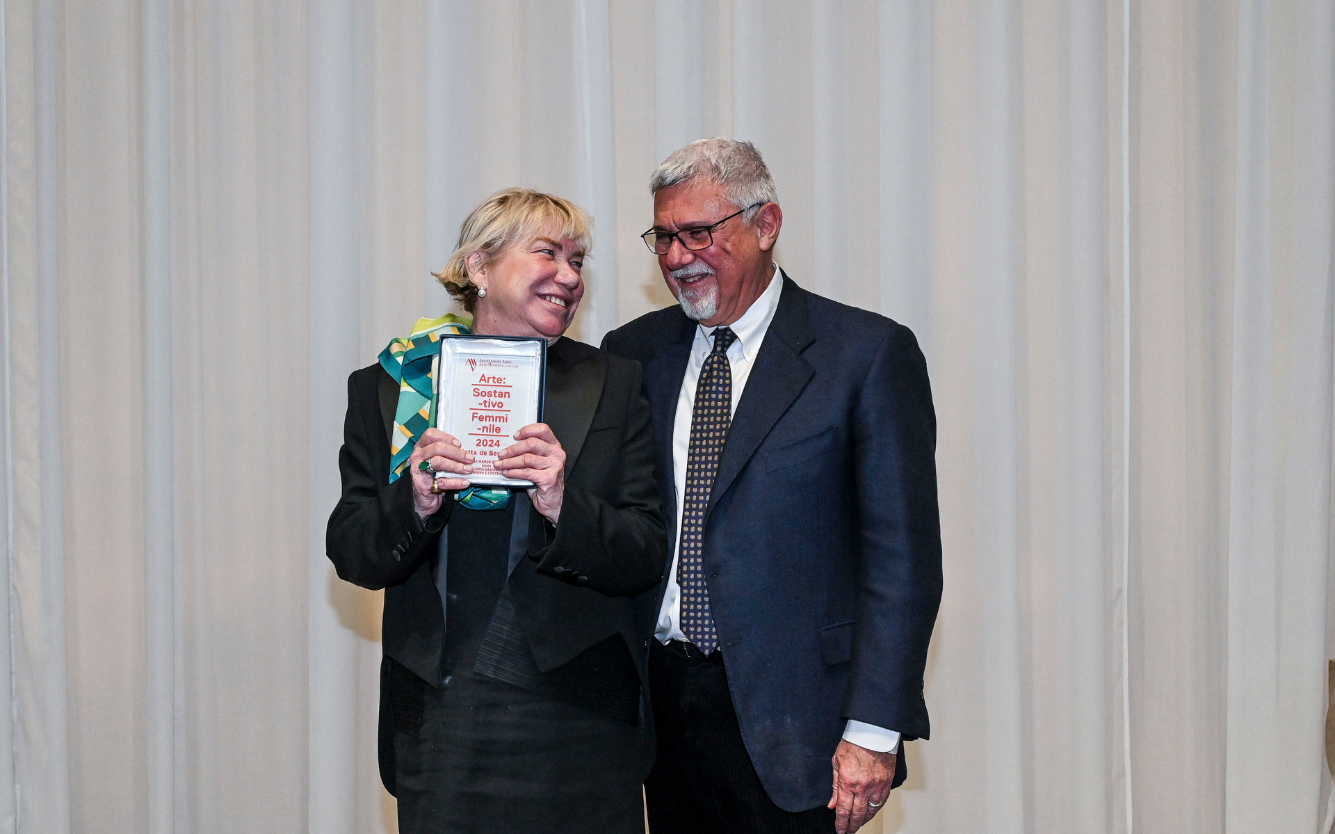 Two people at an award ceremony: a woman holding a plaque and a man in a suit, smiling together on stage.