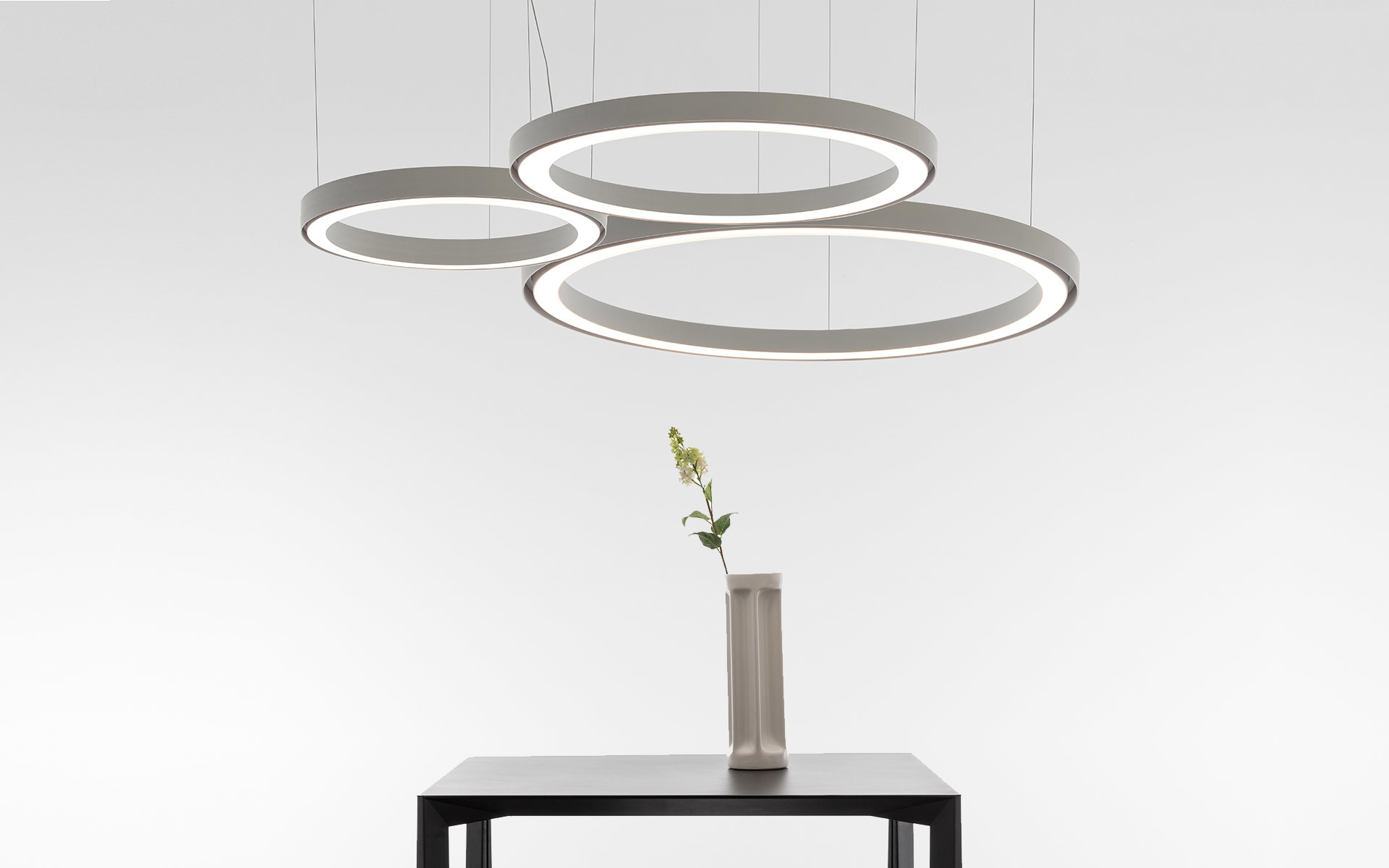Three circular LED pendant lights of varying sizes in grey, suspended above a dark table with a vase and flower.