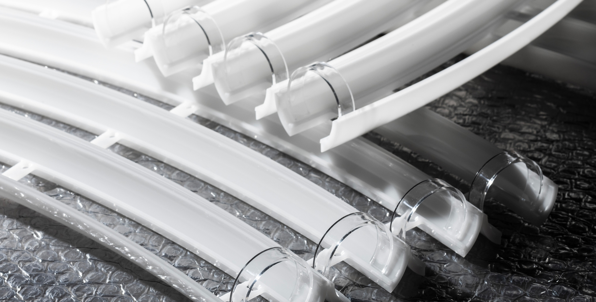 Curved white and transparent extruded profiles for lighting fixtures, arranged on a reflective surface with bubble wrap.