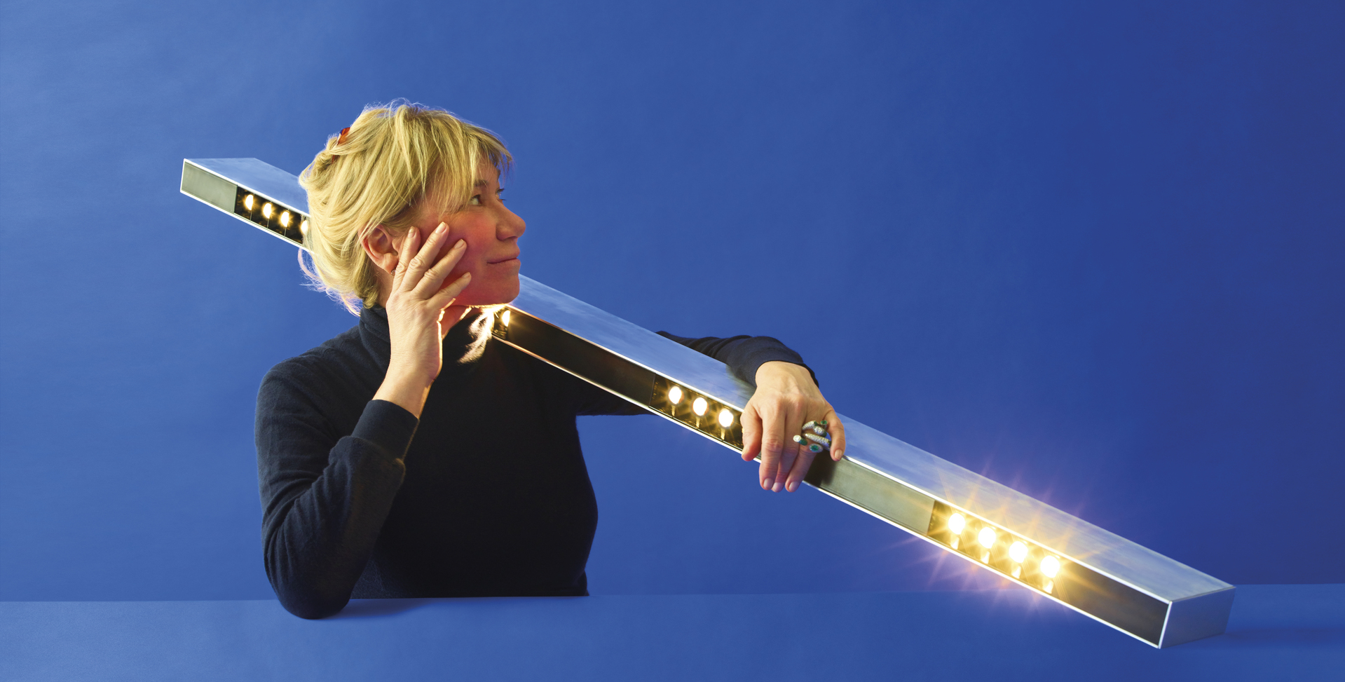 Woman holding an Artemide linear LED bar lamp with warm white spotlights against a blue background.