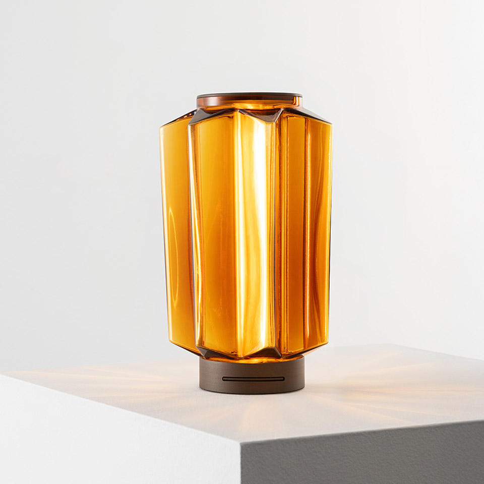 Auralia table lamp by Artemide: faceted amber glass diffuser with bronze base, emitting warm golden light on a white surface