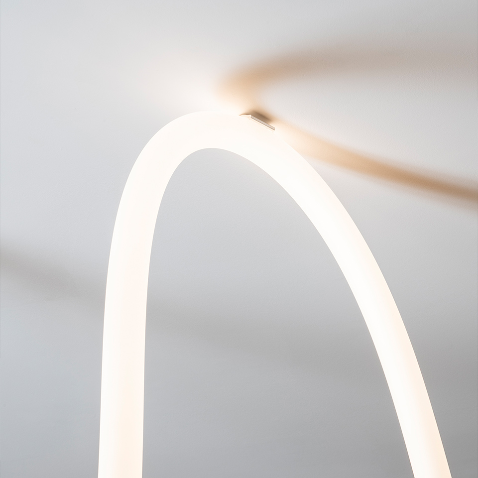 Close-up of Artemide La Linea lamp: a glowing white curved tubular LED arch emitting warm diffused light against a light grey background