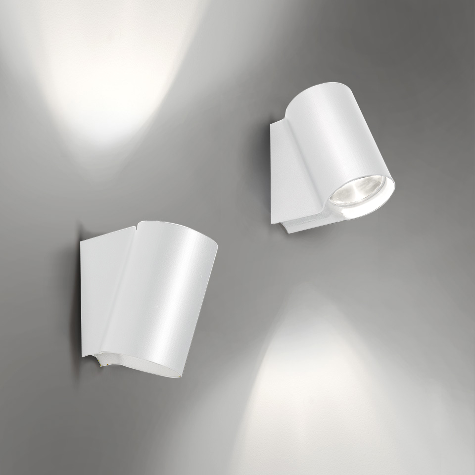 Two Artemide Oblique white wall spotlights: left tilted upward casting light on wall, right angled downward with visible LED source