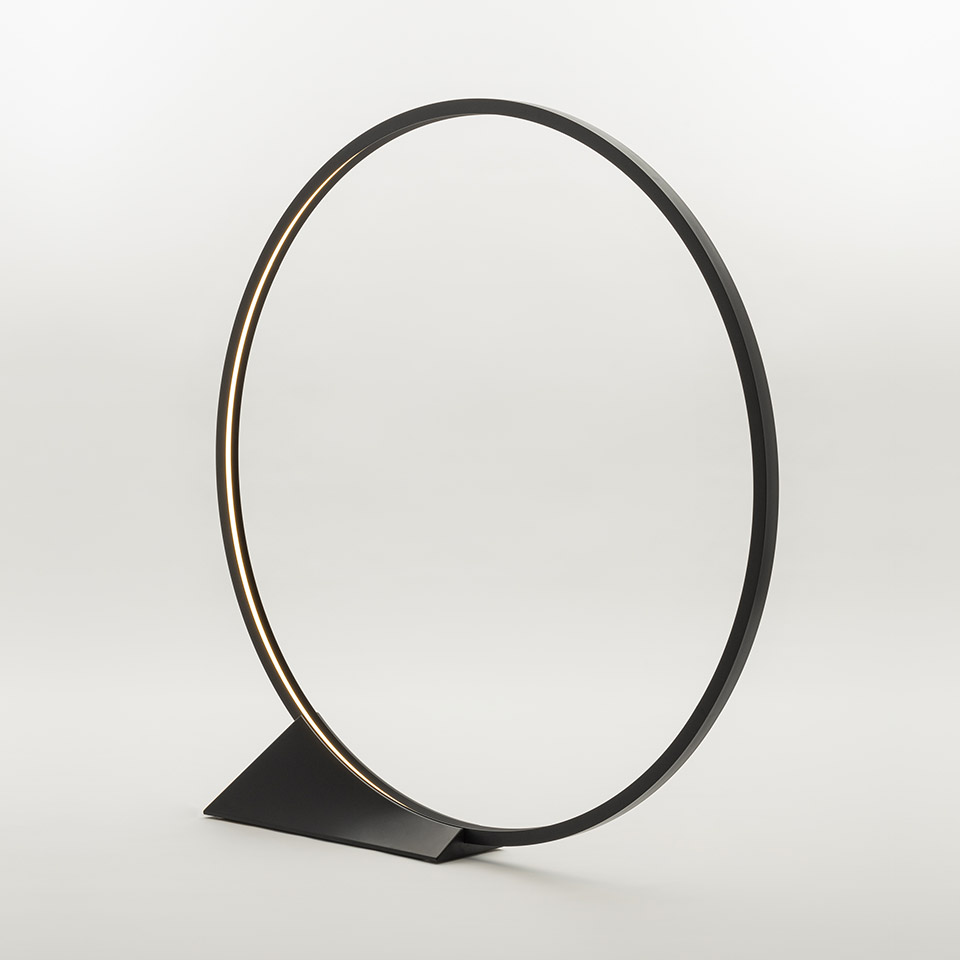 Artemide O table lamp: minimalist black circular LED ring on a flat triangular base, emitting warm light along its inner edge