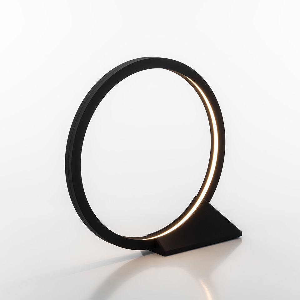 Artemide O table lamp: matte black circular ring with integrated LED strip emitting warm light, mounted on a triangular base