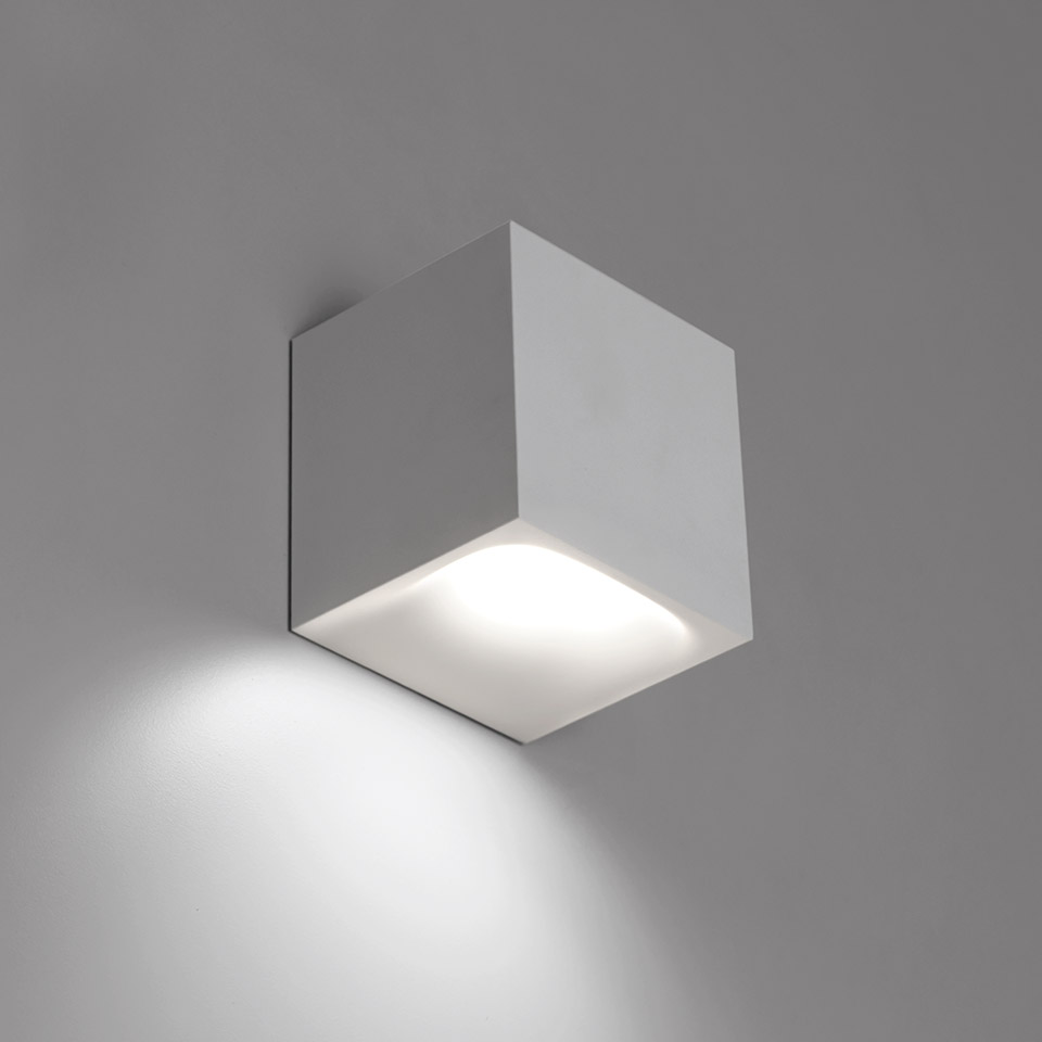 Artemide Aede wall light: compact white cubic body emitting warm downward LED light, mounted on grey wall