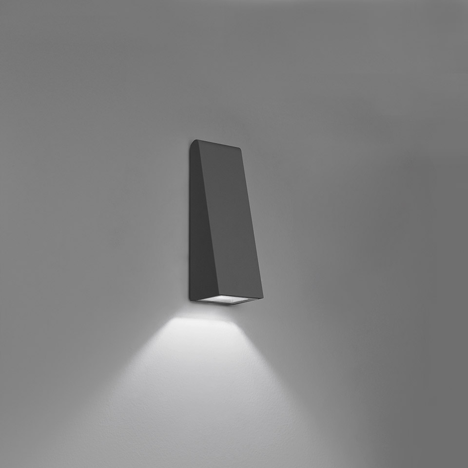 Artemide Cuneo wall lamp, dark anthracite trapezoidal body, emitting focused downward LED light beam on neutral background