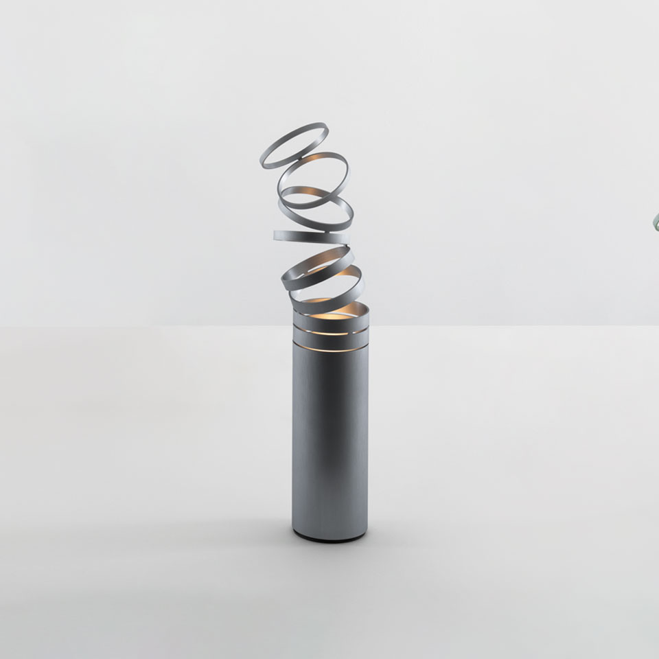 Artemide Decomposé Light Tavolo: cylindrical grey base with an upward spiraling metal ribbon diffuser emitting warm light