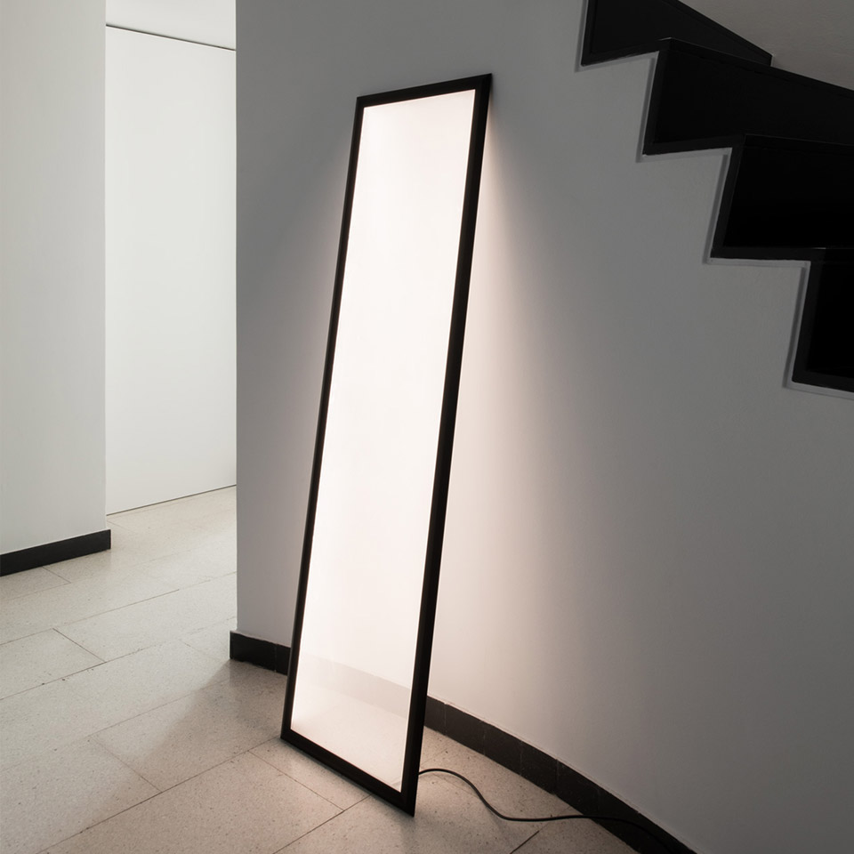 Artemide Discovery Terra floor lamp: tall rectangular luminous panel with black frame, leaning against wall near staircase, emitting soft white light